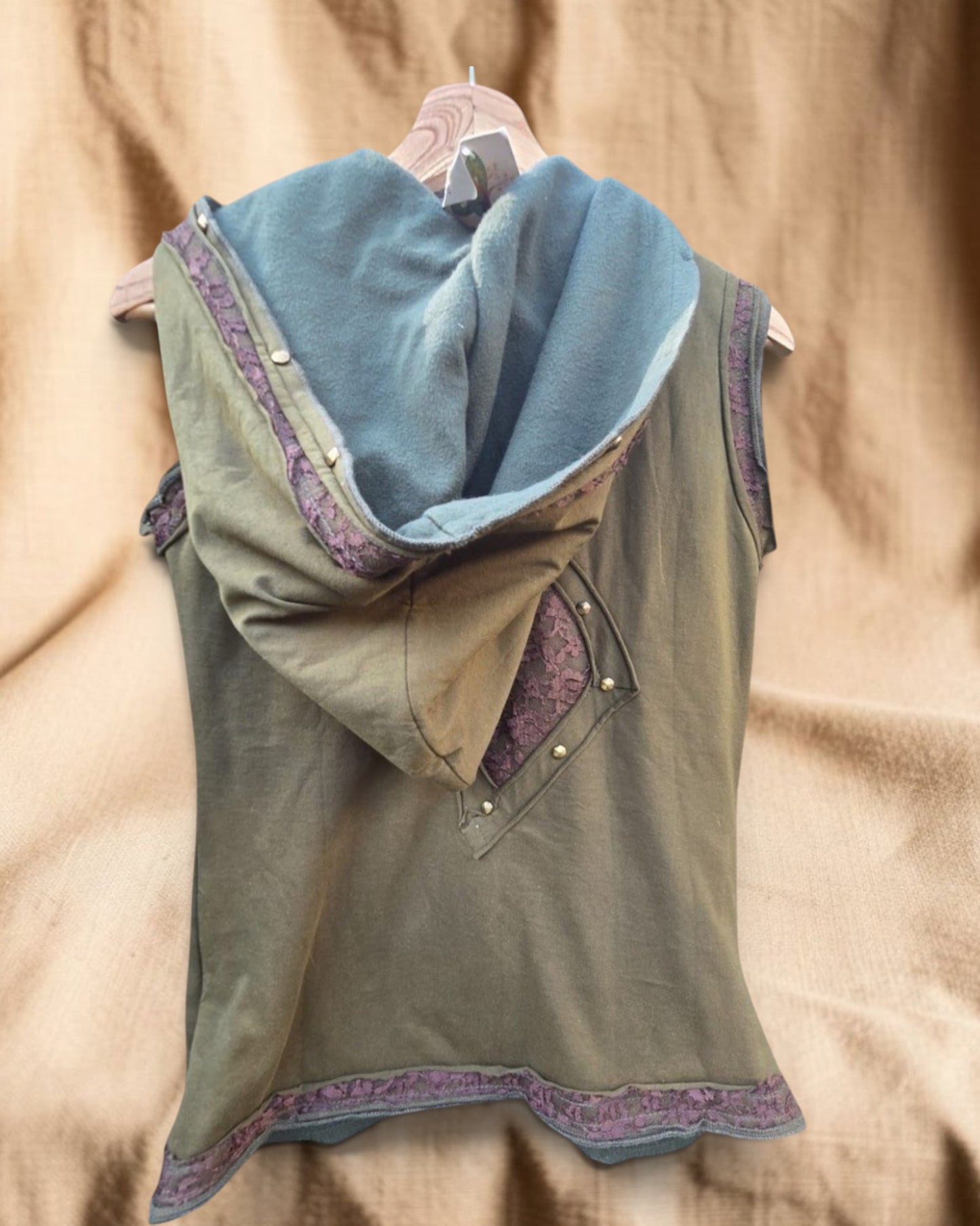 Valentina Sunfade Drift Top, een asymmetrische mouwloze top met capuchon in olijfgroen met paarse kanten details en gouden knopen.