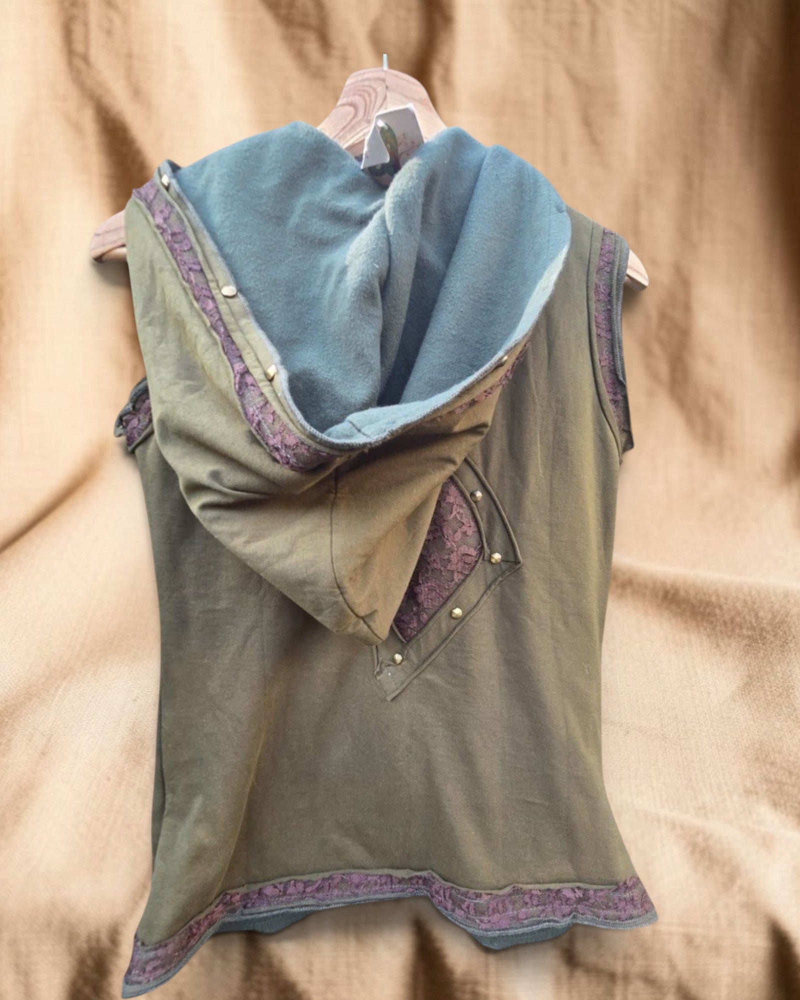 Valentina Sunfade Drift Top, een asymmetrische mouwloze top met capuchon in olijfgroen met paarse kanten details en gouden knopen.
